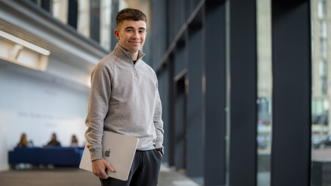 Un estudiante en el pasillo de un edificio moderno sosteniendo una laptop MacBook y sonriendo a la cámara.