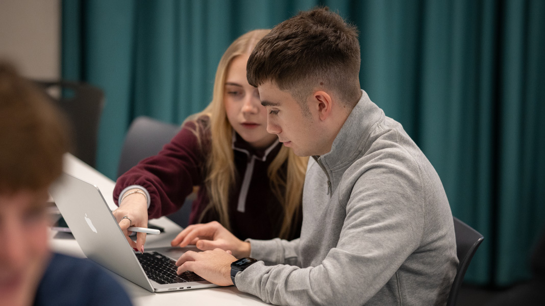 Compañeros de clase colaboran en una MacBook, ella señala la pantalla mientras él escribe