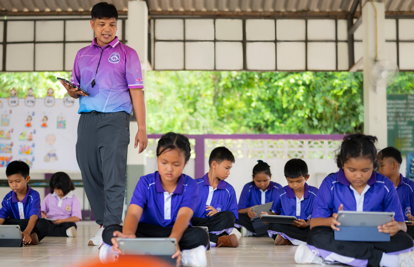 En un aula al aire libre, un profesor de educación física camina entre sus estudiantes, que están en el suelo con sus iPad.