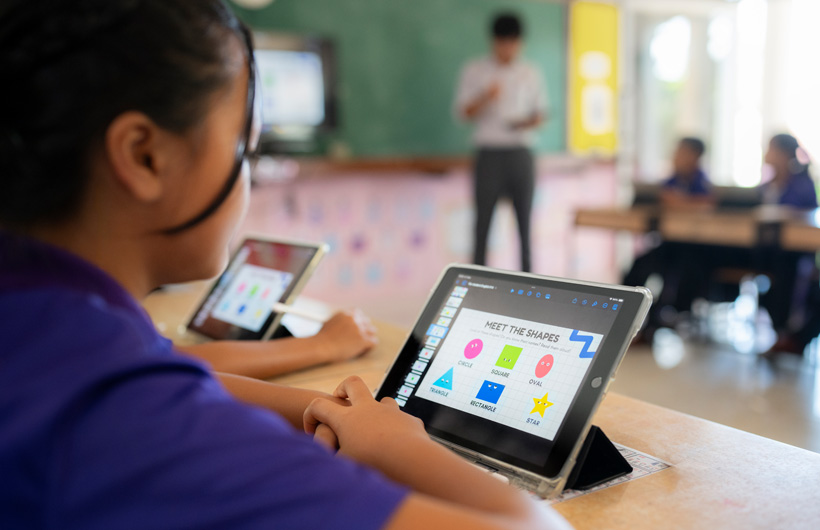 Primer plano de una alumna usando un iPad en su pupitre. En la pantalla se muestra una presentación de figuras geométricas de colores.