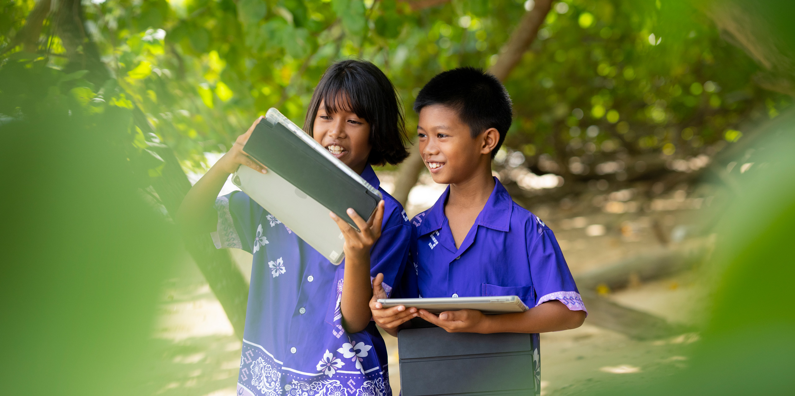 Dos estudiantes de primaria, un niño y una niña, delante de árboles tropicales, cada uno con un iPad. Los dos miran la pantalla del iPad de la niña, sonrientes y motivados.