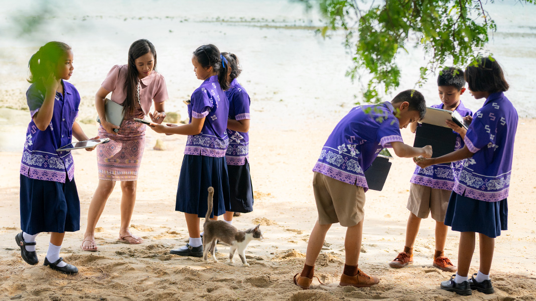 Seis estudiantes de primaria con uniformes morados utilizan sus iPad en una playa de arena. Una profesora ayuda a una alumna con un iPad y hay un gato en medio del grupo.
