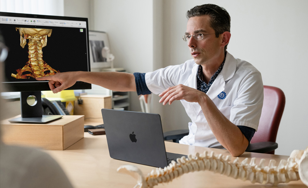 Um médico aponta para uma imagem 3D dos ossos da parte de trás de um pescoço em um Studio Display, e há um MacBook em cima da mesa