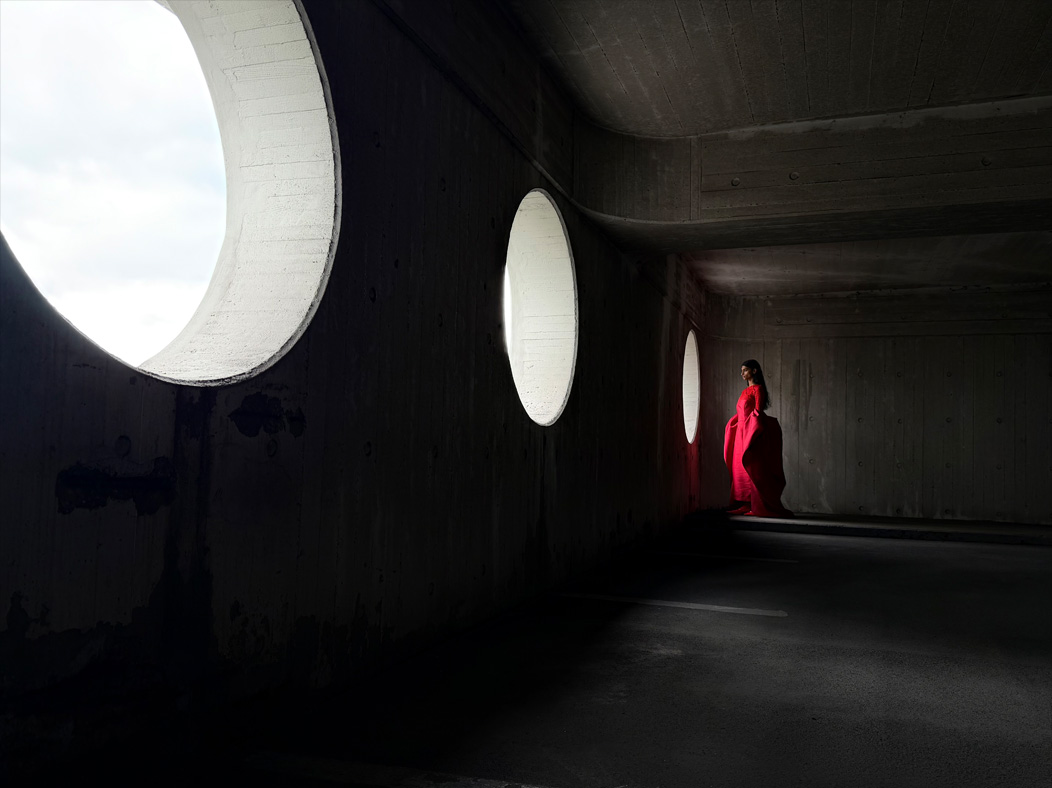 Femme portant une longue robe rouge photographiée dans une pièce vide. L’app Photos montre les distances focales pour illustrer les capacités du système caméra Pro : 28 mm